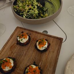 Chopped Salad - Four small appetizers topped with soft-boiled eggs, garnished with chopped green herbs and orange fish roe, arranged on a wooden serving board. The dishes are presented with a drizzle of sauce. In the background, there is a large bowl with a green salad, possibly chopped greens and peas, on a white table surface. The setting appears to be a dining table with utensils and glasses nearby.