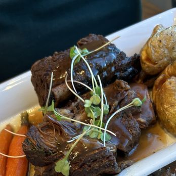Devil's Kitchen - Ribs - A food presentation showing braised beef ribs with a dark glaze, garnished with microgreens. Accompanying the ribs are roasted potatoes and carrots, arranged on a white rectangular plate. The image captures the textures of the meat and vegetables, with a focus on the plated dish. - New American Near Me - Grand Junction, Colorado