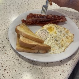 A breakfast plate includes two fried eggs with runny yolks, several slices of cooked bacon, and multiple slices of toasted bread. A person's hand is holding a strip of bacon above the plate, with a fork resting on the plate. The plate is on a speckled, light-colored countertop.