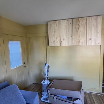 Grand Coast Construction - Hanging cabinets installed - The image shows an interior room with beige walls and a wooden floor. There is a door with a window and a partially installed wooden cabinet above it. An open door reveals a dark hallway. A vacuum cleaner and a cardboard box are on the floor, indicating ongoing construction or renovation. - General Contractors Near Me