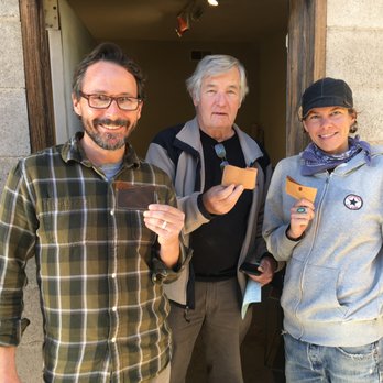 MAKE Santa Fe - The Juki industrial flat bed has been 'hacked' to sew thinner leather. Come learn how to use it! - Three people are positioned outside a building entrance, with a concrete wall and wooden door frame behind them. The man on the left has glasses, a beard, and is wearing a plaid shirt, holding a wallet. The older man in the middle has gray hair, is dressed in a black shirt with a gray jacket, and is holding a cookie and a smartphone. The woman on the right wears a black cap, a gray hoodie with a logo, a blue bandana, and is holding a cookie. All three are smiling at the camera, and the scene appears casual and friendly. - Community Service/Non-Profit Near Me - Santa Fe, New Mexico