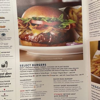 Silver Diner - Menu - A photograph features a Hickory Smokehouse Cheddar Burger with bacon, lettuce, tomato, and pickles on a white plate. The burger has a toasted bun and is accompanied by a side of fries. The background shows a blurred setting, likely inside a restaurant. The menu describes various burger options, including plant-based and chicken alternatives, with additional toppings and substitutions available. - Diners Near Me - McLean, Virginia
