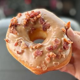 Maple Bacon Donut - A close-up image shows a donut with a light-colored base, topped with a smooth white glaze and sprinkled with small pieces of crispy bacon. The donut is held by a person's hand, with fingers visible on the right side of the image. The background is blurred, emphasizing the donut as the main subject.