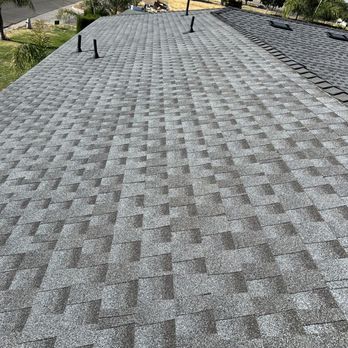Vimahemo Construction - A residential rooftop covered with gray asphalt shingles, featuring several vents protruding from the surface. The roof has a slight slope and is part of a house located in a neighborhood with trees, a street, and parked cars visible in the background. The sky is clear and sunny, indicating daytime. - General Contractors Near Me - Parlier, California