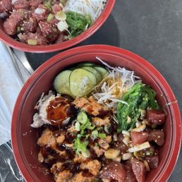 Two red bowls of poke with fresh vegetables, sliced cucumbers, and toppings on a dark table, featuring raw fish, sauces, and garnishes.