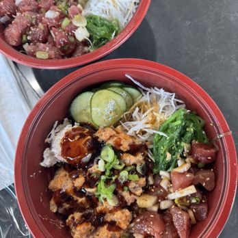 Aloha Cones - Two red bowls of poke with fresh vegetables, sliced cucumbers, and toppings on a dark table, featuring raw fish, sauces, and garnishes. - Poke Near Me - Denver, Colorado