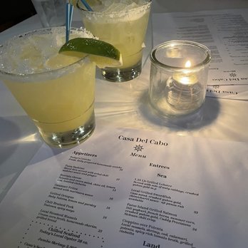 Casa del Cabo - Margaritas - Two margaritas with lime wedges and straws are placed on a table at Casa Del Cabo, a restaurant offering seafood and Latin-inspired cuisine. A candle in a glass holder and a menu are also visible on the table, with a glass of water and a person's arm in the background. - Seafood Near Me - Eastham, Massachusetts