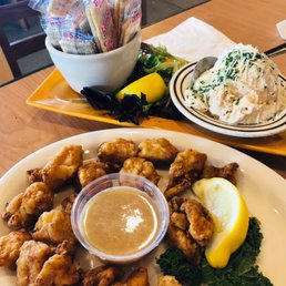 Gator Tail - A plate of fried chicken wings served with a small container of dipping sauce, garnished with a lemon wedge and parsley. Accompanying the main dish is a side of potato salad topped with shredded cheese and herbs, and a side salad with crackers, greens, and a lemon wedge. The food is presented on a white plate and a yellow tray, set on a wooden table in a casual dining setting.