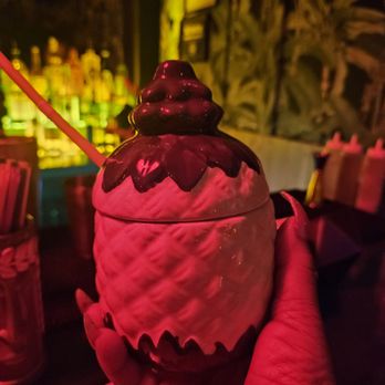 Zig Zag - Painkiller? - A ceramic pineapple-shaped container with a dark chocolate topping is held in a person's hand in a dimly lit bar with colorful bottles in the background. - Cocktail Bars Near Me - Brooklyn, New York