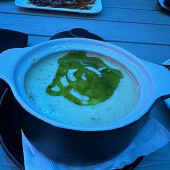Circa69 - Corn and blue crab chowder - A bowl of creamy soup with a green oil drizzle is placed on a black tray on a wooden table at a restaurant, with other dishes visible in the background. - Cocktail Bars Near Me - Jensen Beach, Florida