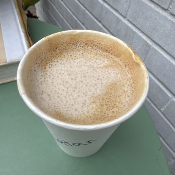 Katan - Pistachio latte - A disposable cup filled with pistachio latte featuring foam on top, set on a green table near a brick wall. - Coffee & Tea Near Me - Queens, New York