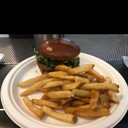 Beyond Burger - A burger with a glossy, toasted bun, fresh leafy greens, and a beef patty is presented on a white disposable plate. The burger is accompanied by a generous serving of French fries, which are golden and slightly crispy. The setting appears to be an airport or casual dining area, with a metal surface and a black background visible behind the plate.