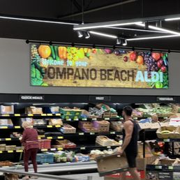 people shopping in a grocery store - The image depicts the interior of an ALDI grocery store in Pompano Beach, FL. Shelves are stocked with various produce and packaged goods, with shoppers browsing. A digital sign overhead reads 'Welcome to your Pompano Beach ALDI,' surrounded by images of fruits and vegetables. The ceiling features modern lighting fixtures, and a shopping cart is visible in the foreground.