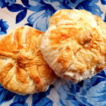 Ruthie's Bagels - Knishes - Two knishes with flaky, golden-brown crusts are positioned on a blue fabric with a floral pattern. The knishes are round, with slightly uneven surfaces and visible layers of pastry, indicating a baked, crispy exterior. The fabric background features dark blue floral designs, providing contrast to the light-colored knishes. - Bagels Near Me - Albuquerque, New Mexico