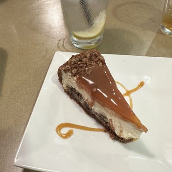 Abingdon Grill - Turtle cheesecake. - A slice of turtle cheesecake featuring a chocolate crust, creamy cheesecake filling, and topped with chopped pecans and a layer of caramel and chocolate glaze. The cheesecake is served on a white square plate with decorative caramel sauce. A tall glass of ice water with a lemon wedge and a straw is positioned behind the plate, on a wooden table surface. - American Near Me - Abingdon, Maryland