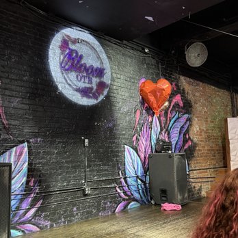 Bloom OTR - The image shows an interior scene of a nightclub or bar with a black brick wall decorated with a colorful butterfly mural in shades of purple, blue, and pink. A projected sign on the wall reads 'Glow on 2,' and a red heart-shaped balloon is attached to the wall. There are speakers positioned on the floor, and the ceiling is black with some peeling paint and exposed wiring. The wooden floor is visible, and part of a person's curly hair is seen in the lower right corner. The setting suggests a lively, decorated space for social gatherings or events. - Pubs Near Me - Cincinnati, Ohio