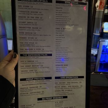 SafeHouse - Menu 10/19/25 - The image shows a menu held by a person at a bar, featuring sections for classified cocktails, base brews, cans & bottles, and other categories. The menu includes drink names, descriptions, and prices, with some items marked as brewed in secret or with special ingredients. The menu is printed on paper with a black border, and the background suggests a bar setting. - Burgers Near Me - Milwaukee, Wisconsin
