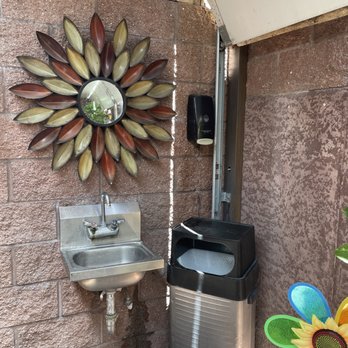 Taco Fish - Look, a sink to wash up - An outdoor setup features a small stainless steel sink, a decorative flower-shaped mirror, and a trash bin, with a brick wall background. - Seafood Near Me - Tucson, Arizona