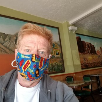 Char's Thai - A person with red hair and blue eyes is taking a selfie indoors, wearing a face mask with comic book-style words like 'Super!' and 'Wow!'. The background features mural paintings of landscapes on the walls, and the room has a ceiling with textured white paint and green walls. There are tables and chairs visible in the background, indicating a casual dining or community space. - Thai Near Me - Tucson, Arizona
