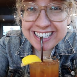A woman with light-colored hair, glasses, and a denim jacket is smiling at the camera. She is holding a cocktail glass with a lemon wedge garnish and a straw, and appears to be in a restaurant or bar setting. The background shows a television screen and dim lighting.
