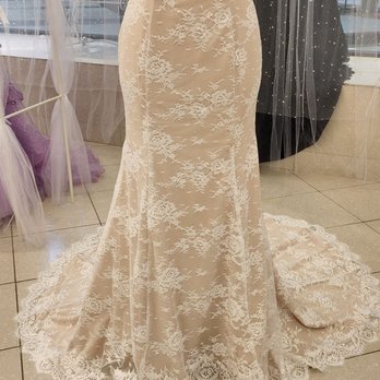 Normandy's Boutique - A beige lace wedding dress with floral embroidery and beaded embellishments is displayed on a mannequin inside a boutique. The dress features a fitted silhouette with lace detailing extending to the train. A matching lace veil and pillow are placed on the floor beside the dress. The boutique has a glass window in the background, with other dresses and accessories visible. - Women's Clothing Near Me - Peoria, Illinois