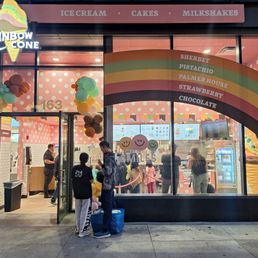 The Rainbow Cone ice cream shop has a large rainbow arch sign with flavors listed, including sherbet, pistachio, Palmer House, strawberry, and chocolate. The storefront is decorated with multicolored balloons, and the interior has pink polka dot walls. Customers are seen inside ordering, and two people are outside, one with a blue bag. The shop offers ice cream, cakes, and milkshakes, and the exterior features a glass window and door.