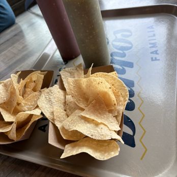 Someburros - Comp chips and salsa - A tray from Someburros in Buckeye, AZ, holds two squeeze bottles of salsa, one red and one green, and two baskets filled with tortilla chips. The tray is on a wooden table, with a blurred background showing part of the restaurant interior. - Mexican Near Me - Buckeye, Arizona