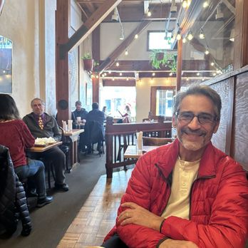 Poppi's Anatolia - So warm and inviting on a cold winter's day. - A man with glasses and a beard, wearing a red jacket over a white shirt, is seated at a wooden table inside a restaurant. The interior features wooden beams, string lights, and large windows allowing natural light. Other patrons are visible in the background, seated at tables along the wall, creating a warm and inviting atmosphere. - Indian Near Me - Eugene, Oregon