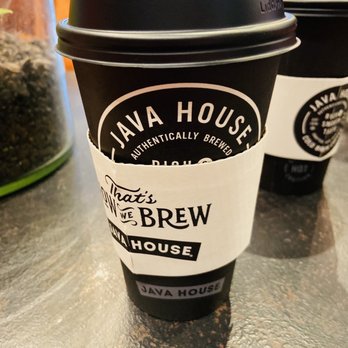 Java House - Chocolate Coconut - A black disposable coffee cup with a white sleeve from Java House, placed on a textured table surface, with another cup partially visible behind it. - Coffee & Tea Near Me - Carmel, Indiana