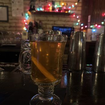Lost & Found - Spiked cider - A glass mug with a handle contains a hot beverage, with a lemon wedge inside, placed on a dark bar counter. Behind the bar, there are metal cocktail shakers, a brick wall decorated with colorful string lights, and bottles on a shelf. The setting appears to be a bar or pub with a cozy atmosphere. - Cocktail Bars Near Me - Washington, DC, District of Columbia