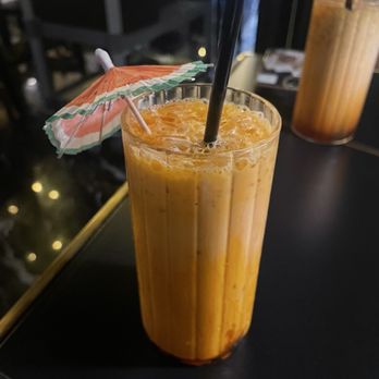 Siam Thai Eatery - Thai Iced Tea - A tall glass filled with Thai iced tea, garnished with a small paper umbrella and a straw, on a dark table. - Thai Near Me - Chicago, Illinois