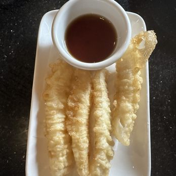 Tsukimi - 2x fried calamari - A white rectangular plate holds several pieces of tempura, which are deep-fried and have a light, crispy batter. A small white cup filled with dark dipping sauce is placed on the plate. The plate is positioned on a black, speckled countertop surface. - Sushi Bars Near Me - Austin, Texas
