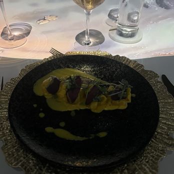 Elysium Immersive Dining - A plated gourmet dish featuring garnishes and sauces is presented on a black plate with a gold decorative rim. The table setting includes a glass of white wine, a glass of water, and additional glasses in the background, all placed on a white tablecloth. The scene suggests a fine dining experience with elegant tableware and presentation. - Modern European Near Me - Lighthouse Point, Florida