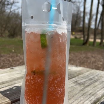 Hop Garden - Blueberry Moscow Mule - DELICIOUS - A blueberry Moscow Mule served in a clear, resealable pouch with a blue straw, placed on a wooden surface outdoors with trees and a cloudy sky in the background. - Breweries Near Me - Paoli, Wisconsin