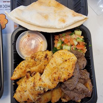 Armo Grill - Chicken and steak, Jerusalem salad, potatoes, and pita bread - The image shows a black plastic takeout container filled with grilled chicken pieces, sliced beef, a cucumber and tomato salad, a folded pita bread on top, and a small container of sauce. The meal appears to be Middle Eastern or Mediterranean cuisine, with a variety of protein, fresh vegetables, and bread, arranged for easy consumption. - Armenian Near Me - Wheeling, Illinois