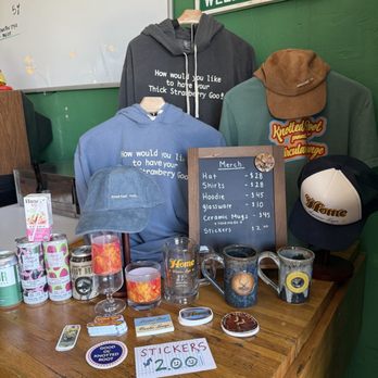Circular Lounge - So much swag! - The image shows a retail display with clothing items, including a gray hoodie, a blue hoodie, and a brown cap, along with ceramic mugs and stickers on a wooden table. A blackboard lists prices for hats, shirts, hoodies, glassware, ceramic mugs, and stickers. Additional branded items like caps and glasses are also visible, with a green wall and a sign in the background indicating a welcoming message for a business related to beer or beverages. - Lounges Near Me - Denver, Colorado