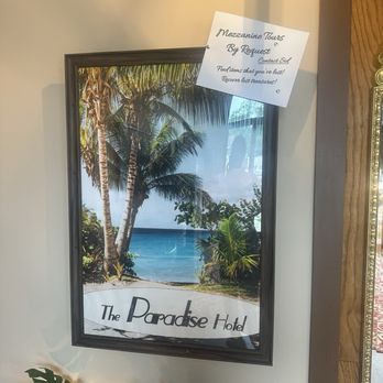 Decode Ann Arbor - A poster of a tropical beach scene with palm trees and ocean, labeled 'The Paradise Hotel,' has a handwritten note attached at the top. - Escape Games Near Me - Ann Arbor, Michigan