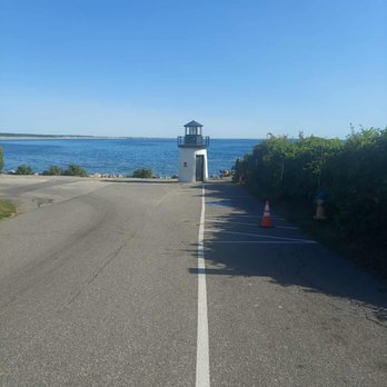 Lobster Point Lighthouse - Lobster Point Lighthouse - A small, white lighthouse with a dark roof is positioned near the shoreline, overlooking a body of water under a clear blue sky. The scene includes a paved area with parking spaces and some traffic cones on the right side, along with bushes and greenery. The water appears calm, and the horizon is visible in the distance. - Landmarks & Historical Buildings Near Me - Ogunquit, Maine