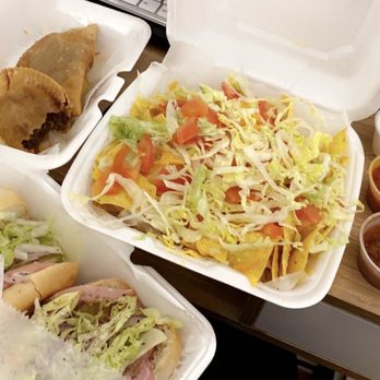 Sofia Pizza House - Italian Sub & Nachos with Chili & Beef Empanadas - Three foam takeout containers are placed on a wooden table, containing tacos with meat and lettuce, nachos topped with shredded lettuce and diced tomatoes, and a sandwich with lettuce, meat, and cheese. Two small cups of red and brown sauces and a small container of sour cream are also visible, along with a computer keyboard in the background. - Italian Near Me - Quincy, Massachusetts