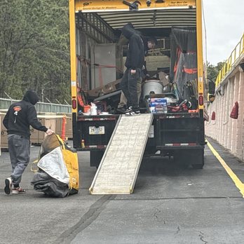 Strongmile Movers - Two people are engaged in loading or unloading items from a yellow moving truck with a ramp. One person is inside the truck, handling items, while the other is outside, pulling a large bag or container. The scene takes place in a parking lot with a tree-lined background and a wall with yellow safety markings. - Movers Near Me - Metuchen, New Jersey