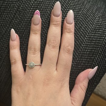Cobblestone Spa & Nails - Almond shaped nails - A hand displays almond-shaped nails painted in a neutral color, with the ring finger featuring pink and purple floral nail art. The hand wears a silver ring with a circular design and is placed on a textured, dark surface. - Nail Salons Near Me - Tempe, Arizona