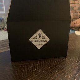 A black cardboard box with a Duchess Cookies logo is placed on a wooden table, with a TV and a brick wall visible in the background.