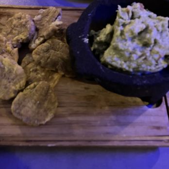 Tiesto's Restaurant - The Tostones and Famous Homemade Guacamole was the perfect compliment before dinner!!! - The image shows a serving of fried plantains and a bowl of guacamole placed on a wooden board. The plantains are sliced and fried, appearing golden brown, while the guacamole is in a black stone bowl, with a creamy texture. The background includes a blue-lit surface and a tray, indicating a dining setting, likely at a restaurant. - Latin American Near Me - Patchogue, New York