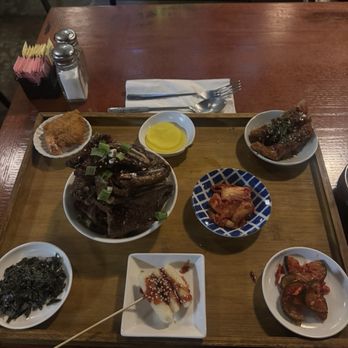Yori - A wooden tray on a restaurant table displays several Korean dishes such as grilled meat garnished with green onions, kimchi, seaweed, and pickled vegetables. Condiments like yellow mustard and dipping sauces are also present. The table has utensils, napkins, and salt and pepper shakers, with a background of a dimly lit restaurant interior. - Korean Near Me - Hurst, Texas