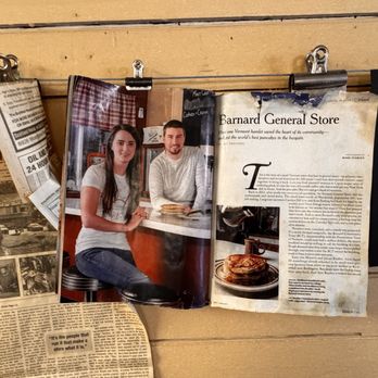 Barnard General Store - History - Multiple photographs and newspaper articles related to Barnard General Store are clipped and hung on a wall. One photo shows two people sitting at a counter inside the store, with a woman and a man smiling at the camera. The articles include text and images about the store's history and community significance. The display is arranged with metal clips on a string, set against a wooden wall. - Grocery Near Me - Barnard, Vermont