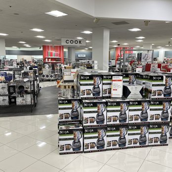 Macy's - Inside - A retail store aisle features a display of Ninja blenders in their boxes, arranged in a pyramid shape. The store has bright lighting, with other kitchen appliances and cookware visible on shelves and racks in the background. Signage indicates different sections, including a cookware area. The store appears spacious and well-organized, with a polished tile floor. - Department Stores Near Me - Concord, California