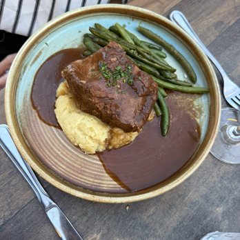 Subourbon - Short ribs - A ceramic plate contains a serving of short ribs topped with gravy and garnished with chopped chives. The plate also includes mashed potatoes and green beans. The setting appears to be a restaurant with a wooden table, and utensils are placed beside the plate. A glass of water and a wine glass are visible nearby. - Southern Near Me - Columbus, Ohio