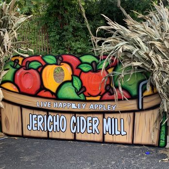 Jericho Cider Mill - a sign for jericho cider mill - The image shows a painted sign for Jericho Cider Mill, depicting a variety of brightly colored apples with green leaves, arranged on a wooden barrel illustration. The sign includes the text "LIVE HAPPELY APPLEY" and "JERICHO CIDER MILL." It is set outdoors, with lush green trees and dried plants visible around the sign, indicating a natural, rustic setting. - Fruits & Veggies Near Me - Jericho, New York