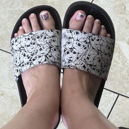 MY TOOOESSS - Feet with decorated toenails in nail art, wearing black slides with a skull pattern, standing on beige tiled flooring.