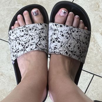 Glitter Box Nails Spa - MY TOOOESSS - Feet with decorated toenails in nail art, wearing black slides with a skull pattern, standing on beige tiled flooring. - Skin Care Near Me - Mesa, Arizona